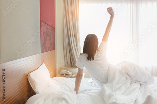 Woman stretching in bed after waking up, back view. Woman sitting near the big white window while stretching on bed after waking up with sunrise at morning, back view.