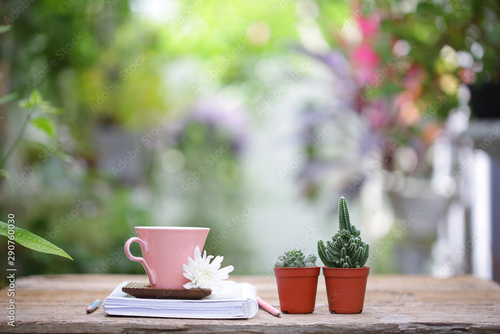 Pink coffee cup with small cactus plant in brown pot and flowers with diary notebooks on wooden table 