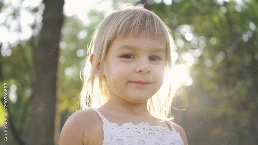 Little gloomy girl looking at the camera and then smiling. Mood changing. Portrait of cute child in the park in sun rays Little gloomy girl looking at the camera and then smiling. Mood changing. Portrait of cute child in the park in sun rays