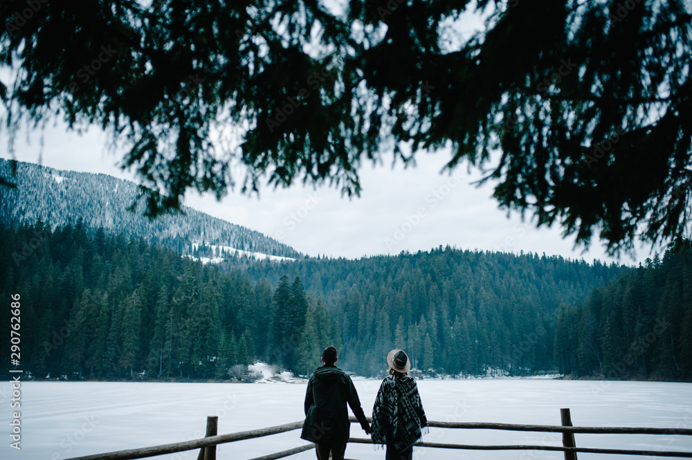 Young couple back standing on pier near the lake on nature. Winter lake against the background of the forest and the mountains. Landscape. Place for text and design. Close up.