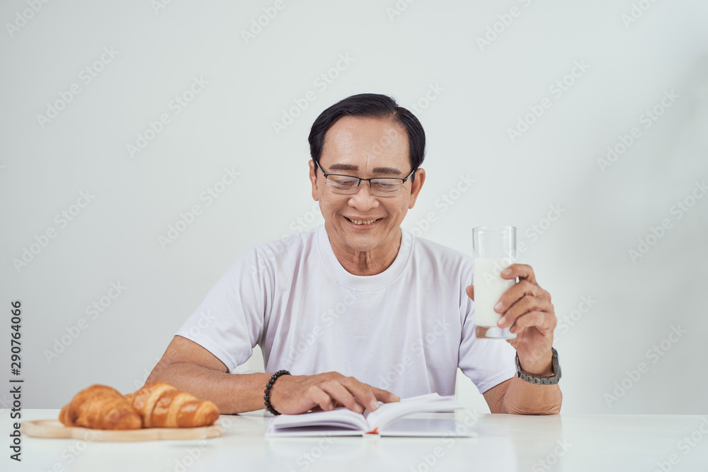 Elder man drinking milk and reading book while having breakfast Elder man drinking milk and reading book while having breakfast