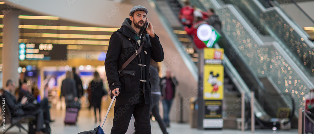 Fototapeta premium Young handsome traveler walking in modern airport terminal, using smartphone app in public wifi area, messaging, travelling with luggage bag, wearing casual style clothes