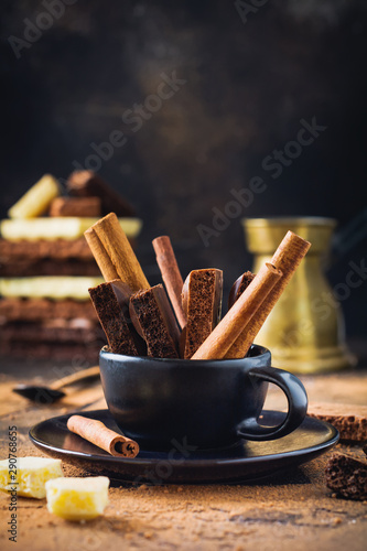 Pieces of porous chocolate and cinnamon sticks in black coffee cup on dark old background. Selective focus.