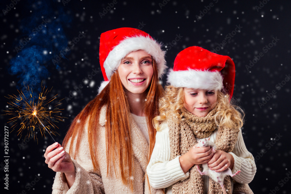 redhaired ginger woman in warm sheep's wool coat and santa claus hat and little blonde girl wear knitted scarf holding bengal sparkler and white rat in studio black background redhaired ginger woman in warm sheep's wool coat and santa claus hat and little blonde girl wear knitted scarf holding bengal sparkler and white rat in studio black background