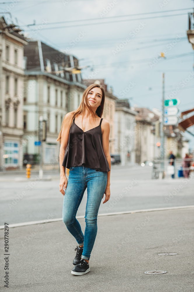 Outdoor portrait of beautiful young woman walking down the road in the city, wearing black cami top and denim jeans. Image taken in Lausanne downtown, place Bel Air, Switzerland