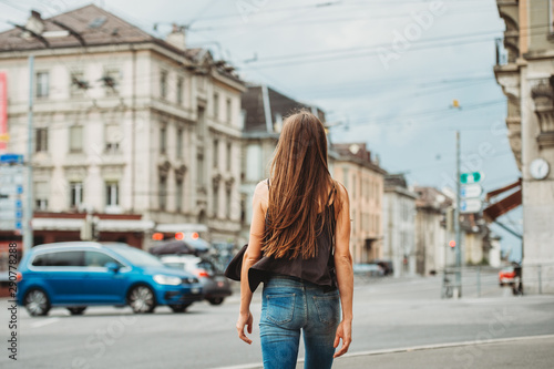 Outdoor portrait of beautiful young woman walking down the road in the city, wearing black cami top and denim jeans. Back view. Image taken in Lausanne downtown, place Bel Air, Switzerland