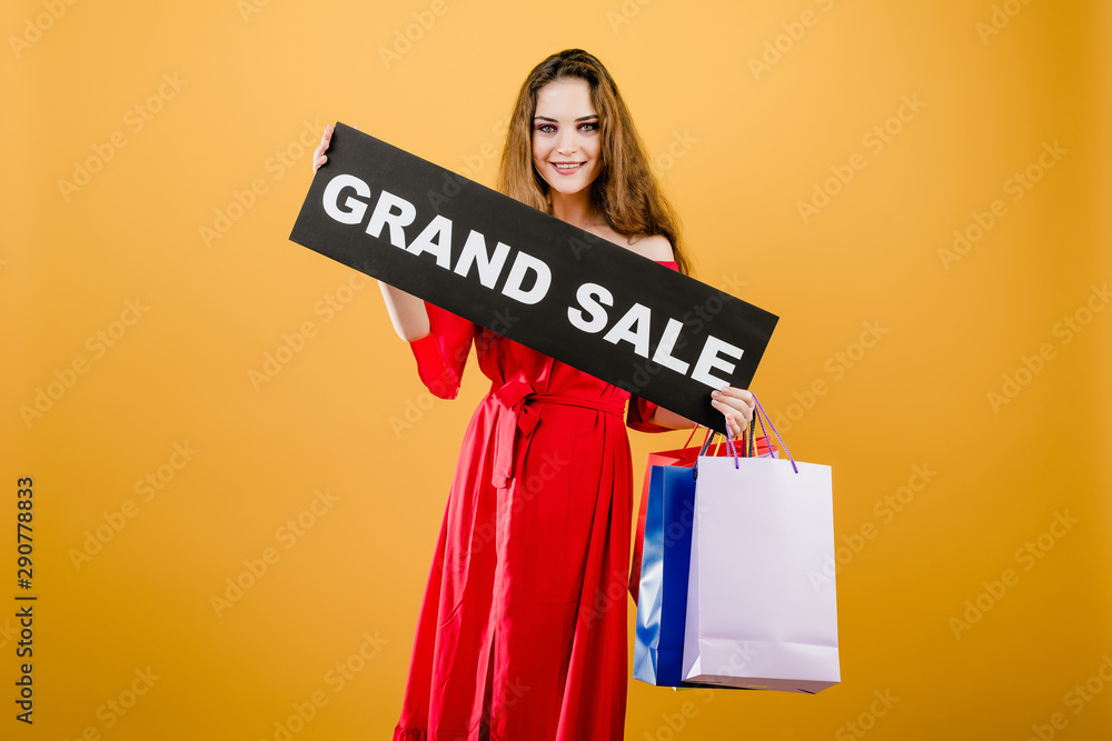 smiling young woman has grand sale sign with colorful shopping bags isolated over yellow smiling young woman has grand sale sign with colorful shopping bags isolated over yellow