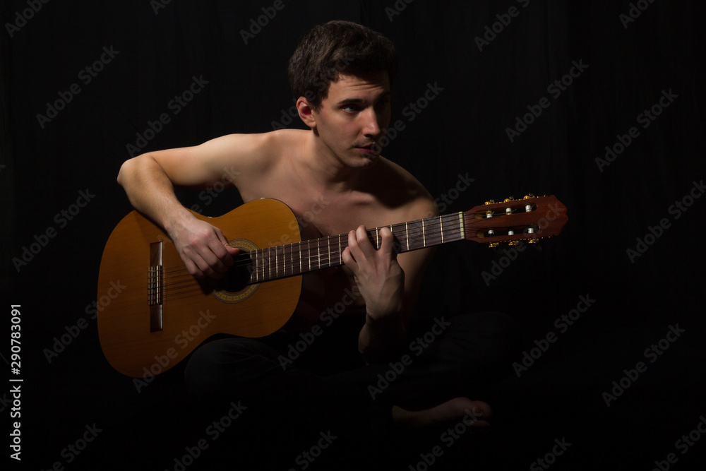 Fototapeta premium Handsome young musician playing on guitar. Naked torso. On black background.