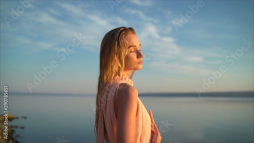 Beautiful young woman standing in sea with waves on sandy beach against orange sky at sunset. Summer travel. A young girl is standing against the sunset in a summer dress.