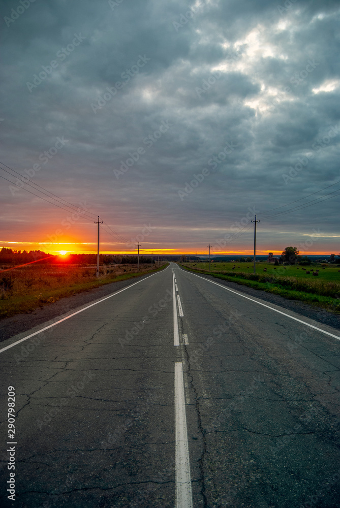 Beautiful nature landscape. The sun sets over the horizon, the last rays. Asphalt country road. Dark clouds.