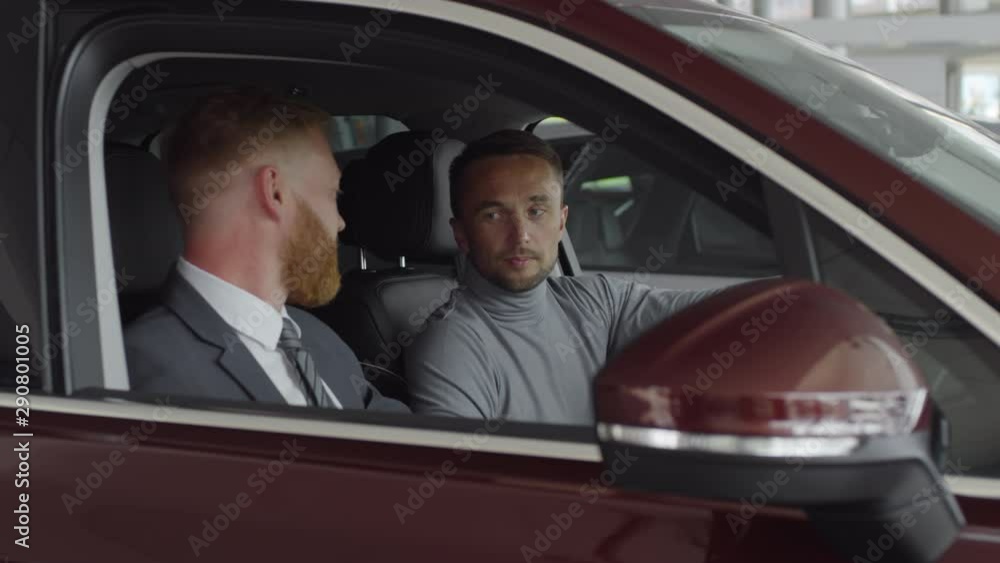 Man sitting in driver seat of car and speaking with professional salesman while choosing new auto in dealership showroom Man sitting in driver seat of car and speaking with professional salesman while choosing new auto in dealership showroom