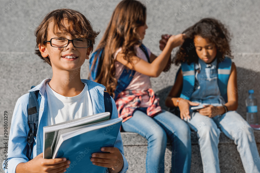 Portrait shot of little school boy in eyeglasses smiling and looking at camera with classmates on background outdoors