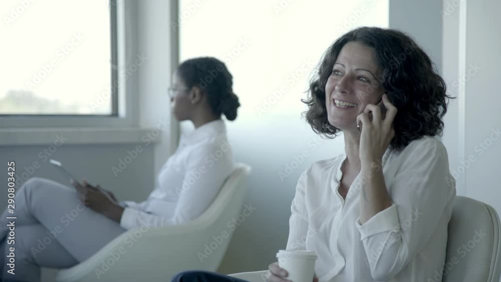 Happy businesswoman talking by smartphone. Cheerful woman drinking from paper cup and talking by cell phone while female colleague using tablet pc behind in office. Technology concept Happy businesswoman talking by smartphone. Cheerful woman drinking from paper cup and talking by cell phone while female colleague using tablet pc behind in office. Technology concept