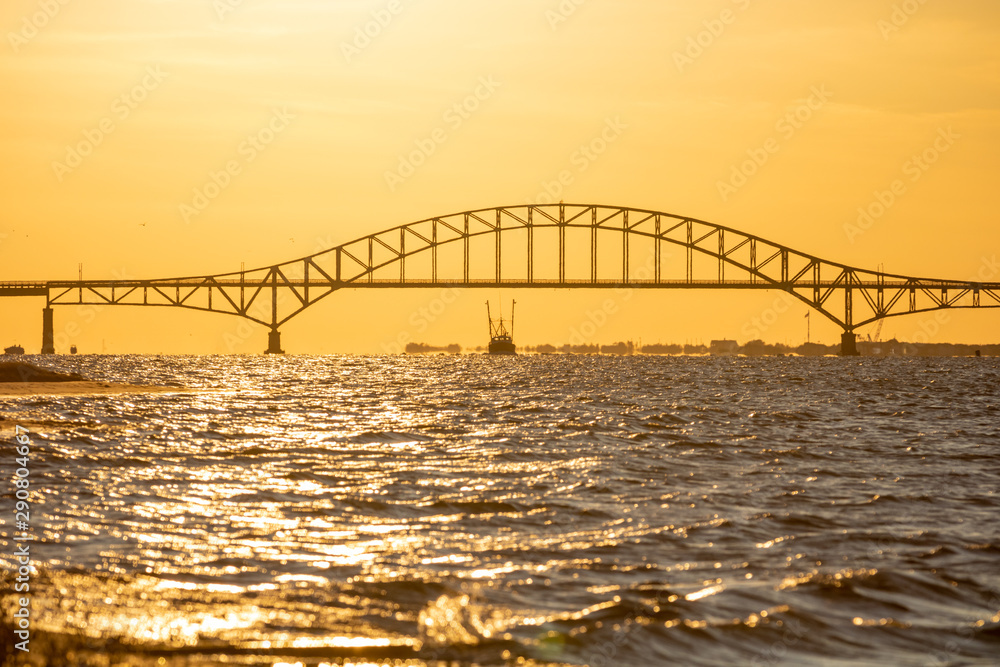 Fishing boat sailing under a steel tied arch span bridge during sunset, with golden light shining over the water. Great South Bay, Long Island New York. Fishing boat sailing under a steel tied arch span bridge during sunset, with golden light shining over the water. Great South Bay, Long Island New York.
