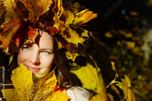 Portrait of a young brunette girl with blue eyes in a wreath of autumn maple leaves on a background of yellow and red foliage