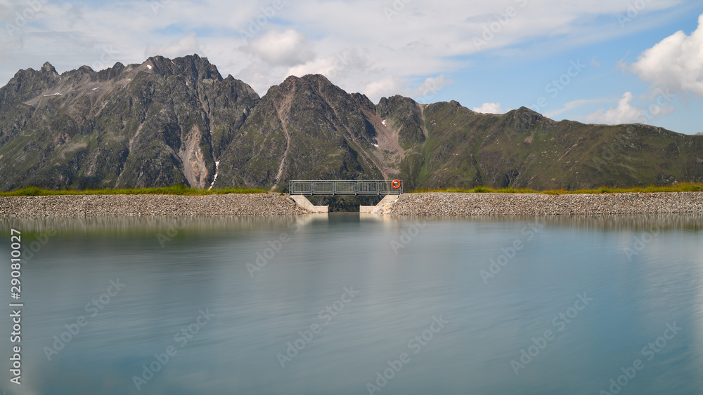 Smooth waters of an alpine water reservoir high up in the Tyrolean mountains in Austria. A modern footbridge with attached red lifebelt crosses the lake. 
