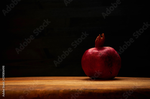 Dark red pomegranate on textured medium gold tone wooden chopping board against dark wooden background. Soft warm light from above, front view. Punica granatum