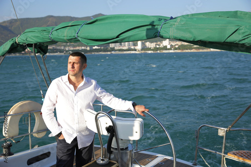 attractive man in white stands at the helm of a yacht at sea. Sailing, tourism, travel and people concept. Happy man at the on steering wheel and navigation.