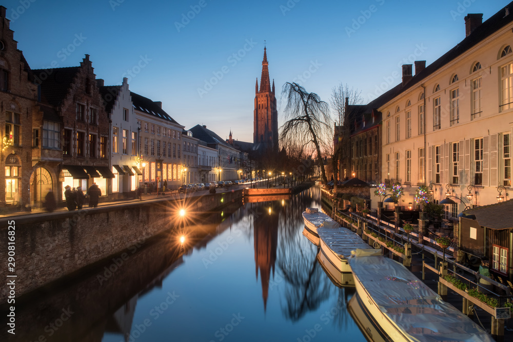 Church of our lady  with traditional building facade reflects on still canal with tourist boats foreground during twilight at Bruges, Belgium