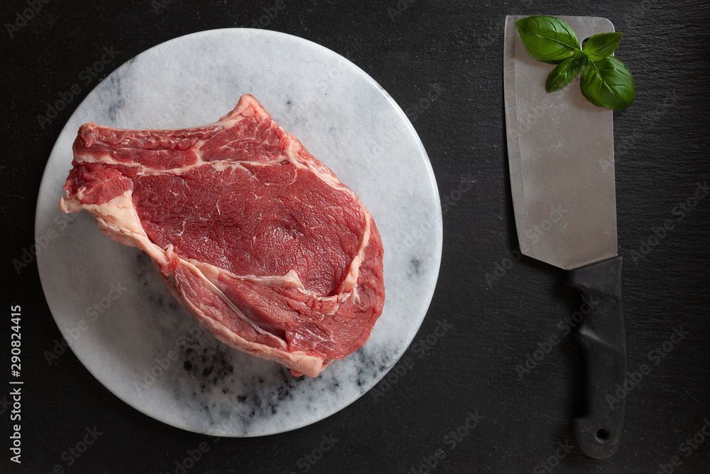 Raw fresh Beef steak with bone, spices and seasonings on stone cutting board. Top view, close upon black background