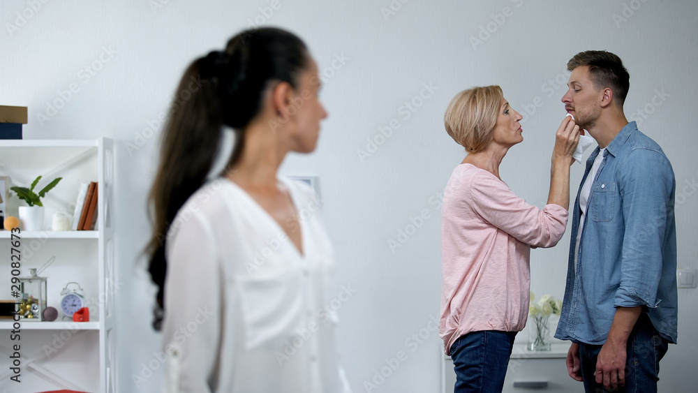 Wife looking at mother-in-law wiping husbands mouth, overprotection problem Wife looking at mother-in-law wiping husbands mouth, overprotection problem