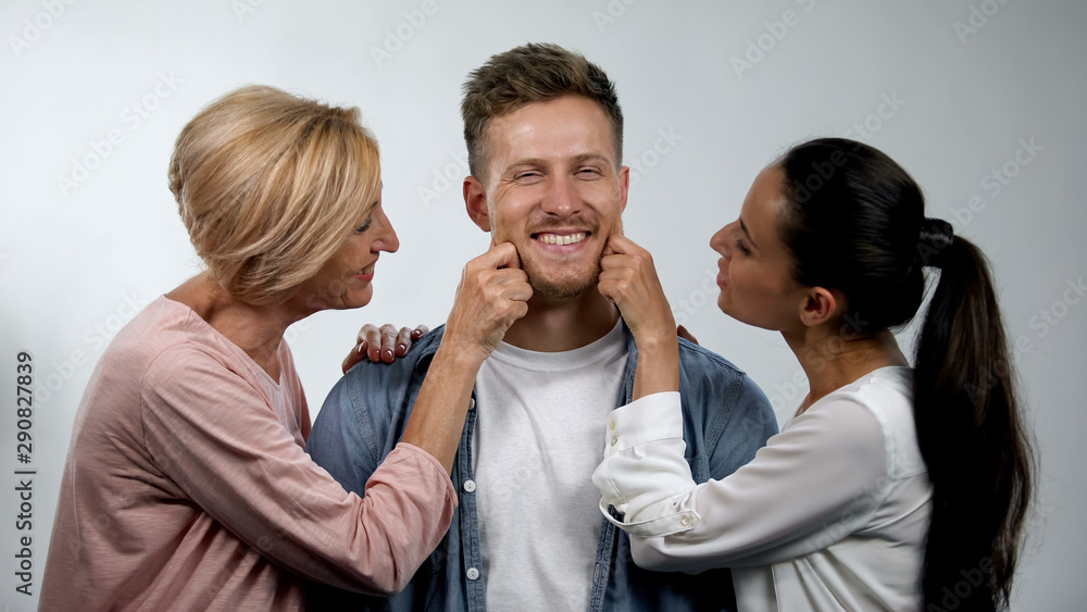 Wife and senior mother pinching young man cheeks, overprotection in family Wife and senior mother pinching young man cheeks, overprotection in family