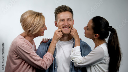Wife and senior mother pinching young man cheeks, overprotection in family