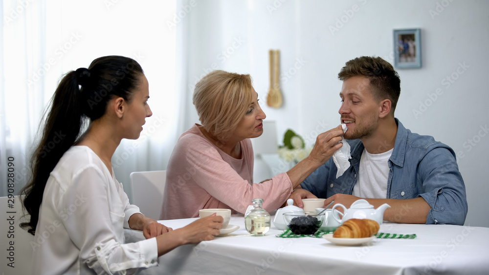 Shocked wife looking at mother-in-law wiping husbands mouth during tee party Shocked wife looking at mother-in-law wiping husbands mouth during tee party