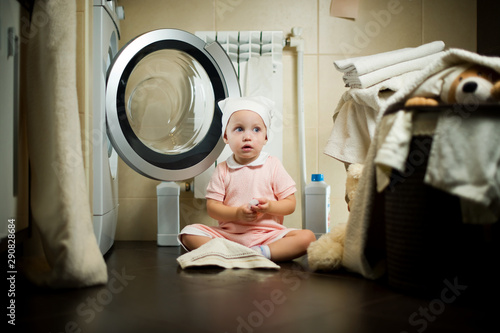 A little girl sits near the washing machine on the floor in the bathroom. In front of her is a towel  a pool of water and many different linen around. The child looks askance at a stack of towels