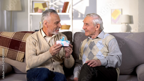Smiling old male holding birthday gift sitting with friend on sofa, celebration