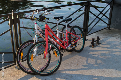 In the morning two bikes white and red of travelling together persons parked and locked with anti-theft cables to the fence street on the background of azure water in the river