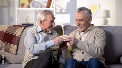 Aged man presenting birthday cake with candle to smiling friend, anniversary