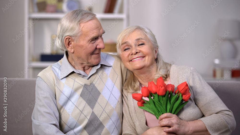 Elderly couple hugging at home, woman holding bunch of tulips, anniversary gift - obrazy, fototapety, plakaty 