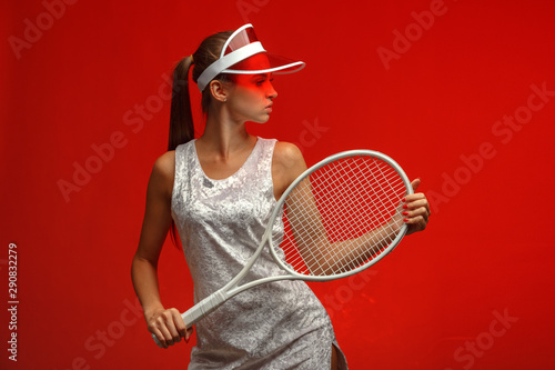 girl tennis player in a white dress in a sun visor with a tennis racket. Red background. Studio photography. © Max
