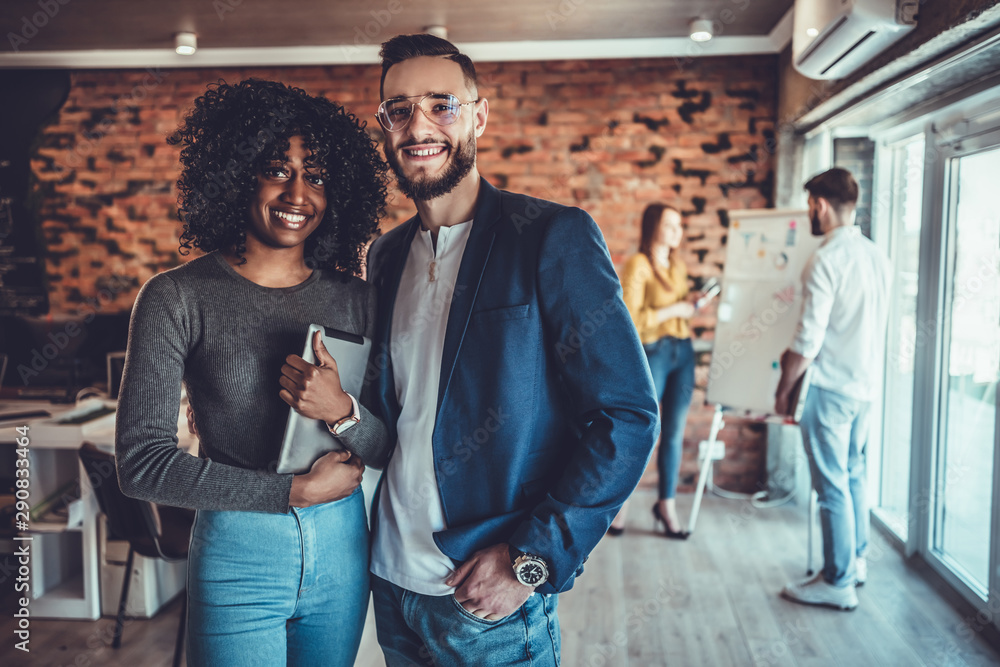 Portrait of young man and woman standing in office with colleagues meeting in background. Portrait of young man and woman standing in office with colleagues meeting in background.