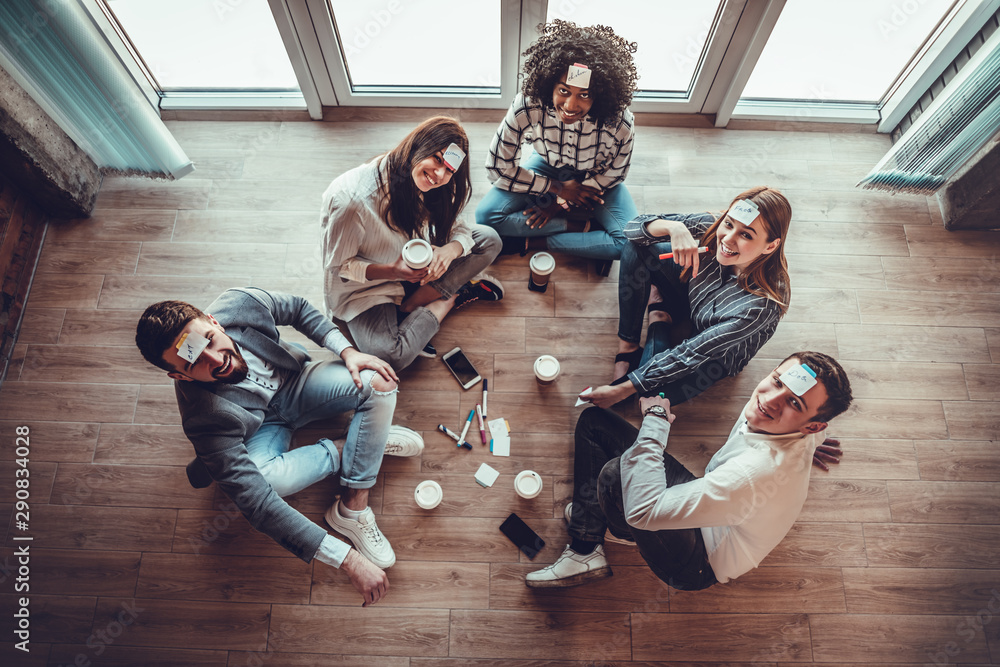 Fototapeta premium Group of young people in office floor having fun playing name game with sticky notes to their forehead