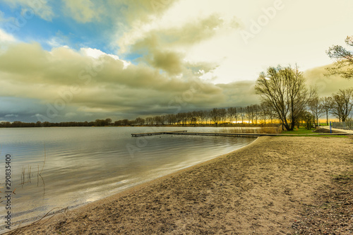 Sunrise at freshwater recreational lake Zegerplas Alphen aan den Rijn with a view of the sandy beach with T-jetty against heavily cloudy skies and breaking sun on the horizon