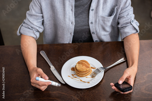 overhead view of woman holding blood lancet and glucose monitor near plate with tasty pancakes