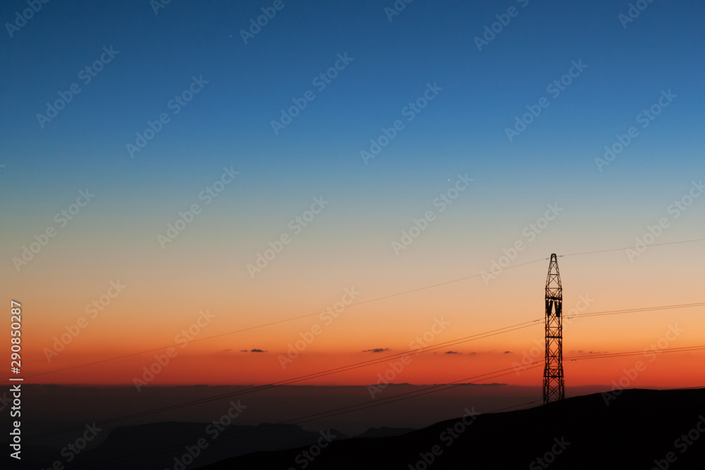 Photograph of a sunset over the mountains in Jordan, with a pole for electricity cables. Beautiful orange, blue, light blue and yellow colors.