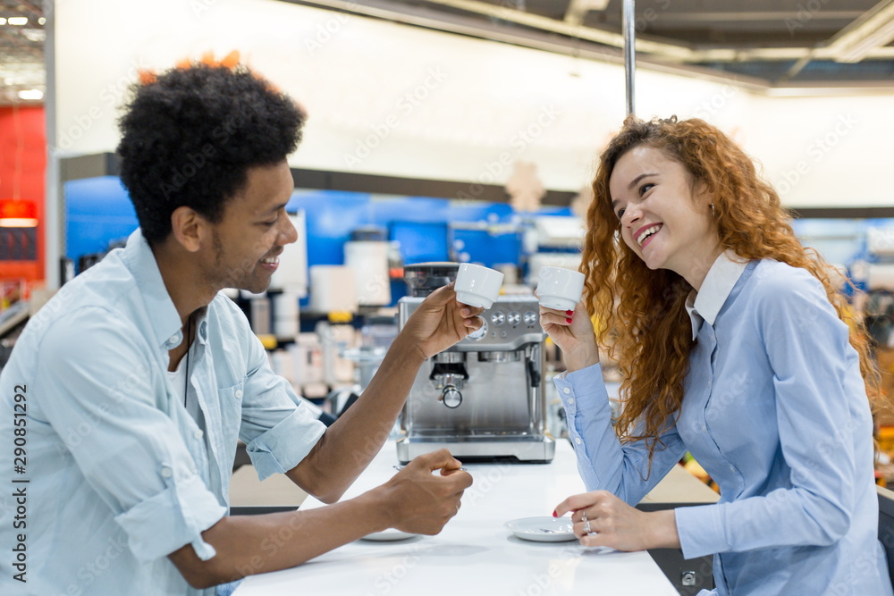 African curly-haired man with a red-haired girl drinking freshly brewed coffee testing coffee machines in the store