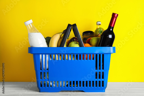 Shopping basket with grocery products on white wooden table against yellow background