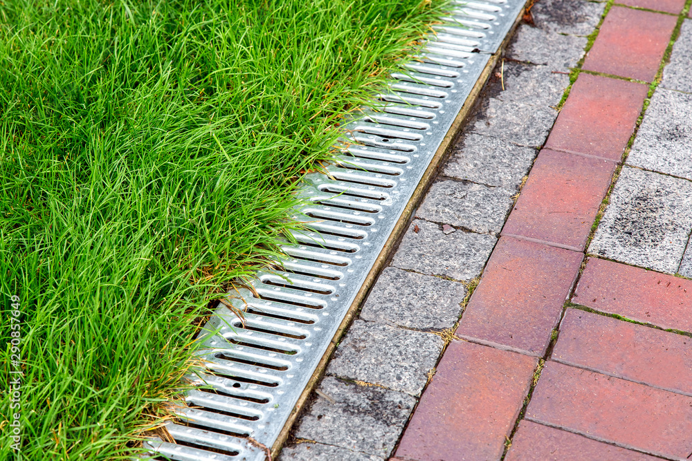 An iron gutter with grate to the drainage system on the side of the footpath with green lawn, close up.
