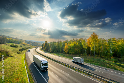 Obraz na płótnie Trucks driving on the asphalt highway between deciduous forest in autumn colors under the radiant sun and dramatic clouds. View from above. w salonie