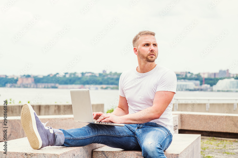 Young American Man with beard, traveling, working in New York City, wearing white T shirt, blue jeans, sneakers, sitting on stone bench at park by river, working on laptop computer, looking, thinking