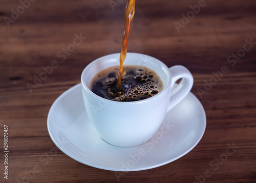 Coffee pouring down in white cup  close up view. White coffee cup and saucer on brown wooden table. Foaming coffee in cup. Selective soft focus. Blurred background