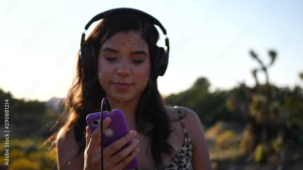 A beautiful young hispanic woman holding a smartphone listening to happy music on headphones outdoors in epic sunlight with lens flares in slow motion. A beautiful young hispanic woman holding a smartphone listening to happy music on headphones outdoors in epic sunlight with lens flares in slow motion.