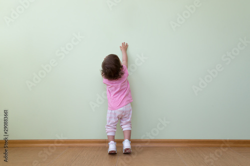 Adorable baby girl in a pink blouse and striped pants stands in front of a green wall and looks up.
