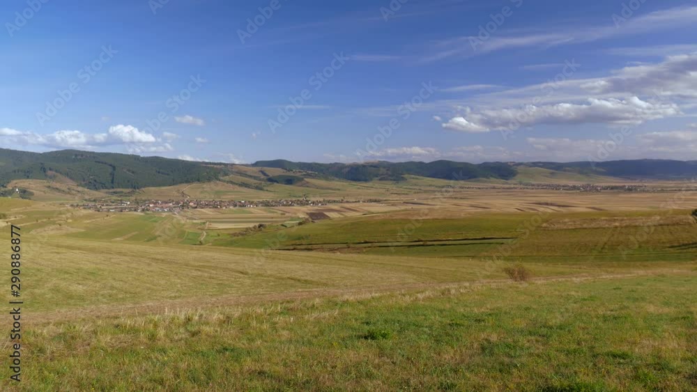 Empty agricultural field, small village in the valley, blue sky with white clouds in Transylvania, Romania.