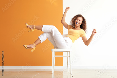 Fashionable young African-American woman sitting on chair against color wall