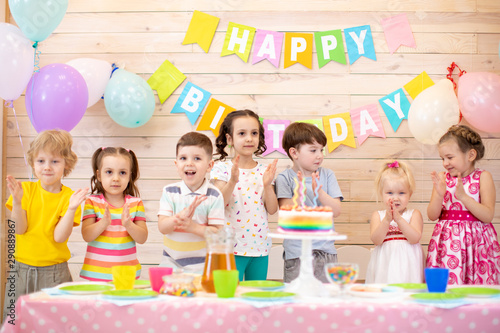 Group of preschool kids clap their hands near birthday cake, balloons on background